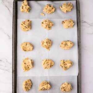 A baking sheet lined with parchment paper holds twelve scoops of raw cookie dough, spaced evenly in three rows.