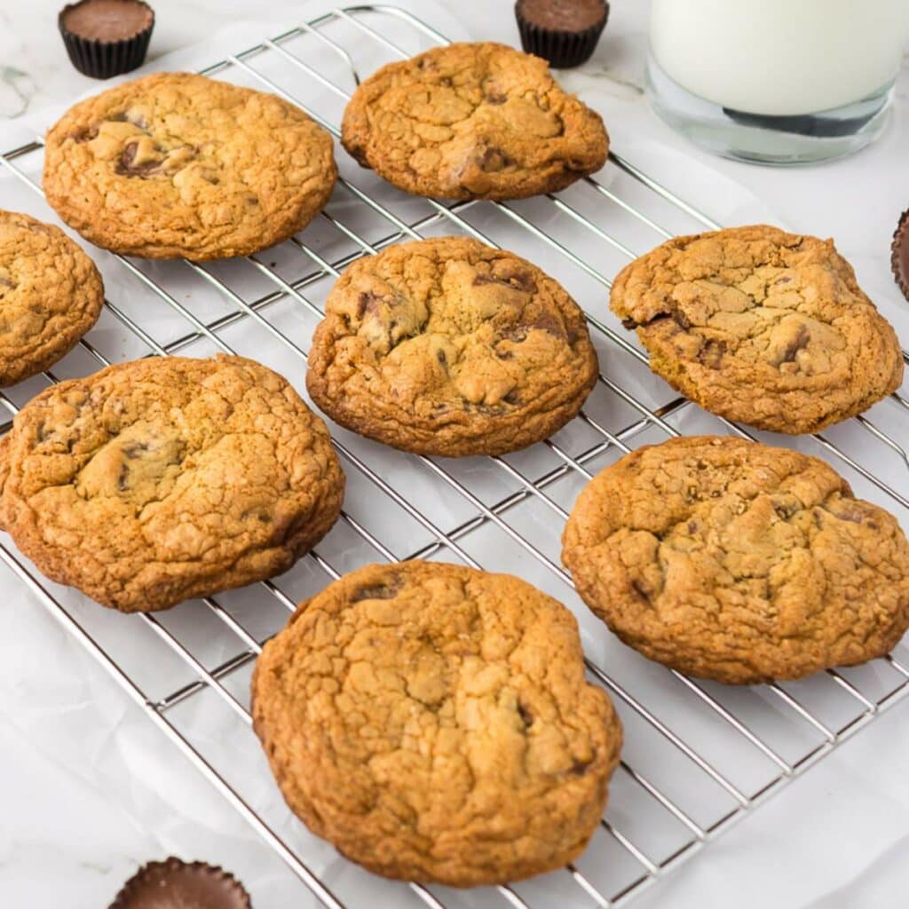 Eight chocolate chip cookies cooling on a metal rack, with a glass of milk and small chocolate candies in the background.