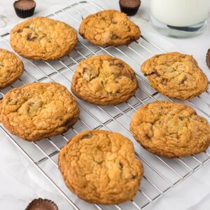 Eight chocolate chip cookies cooling on a metal rack, with a glass of milk and small chocolate candies in the background.