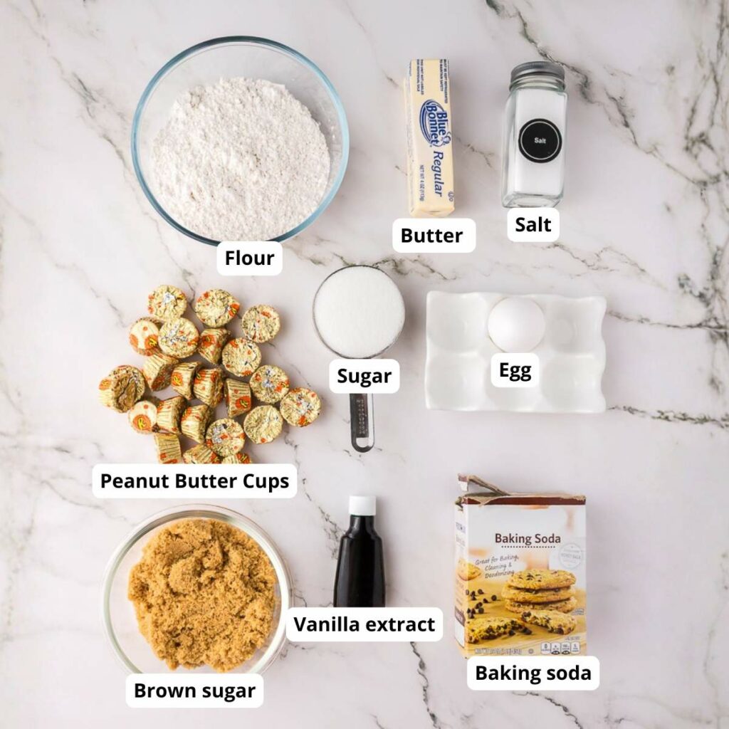 A marble countertop displays labeled baking ingredients: flour, butter, salt, sugar, peanut butter cups, egg, brown sugar, vanilla extract, and baking soda.