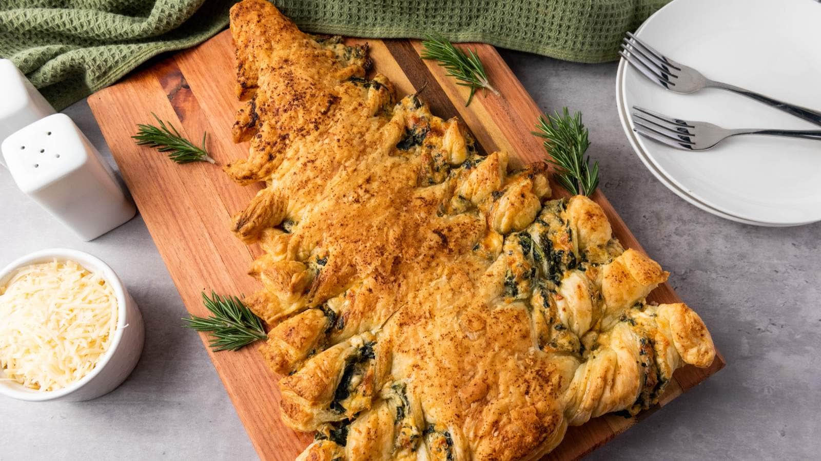 Puff pastry shaped like a Christmas tree, filled with spinach and cheese, on a wooden board with rosemary garnish.