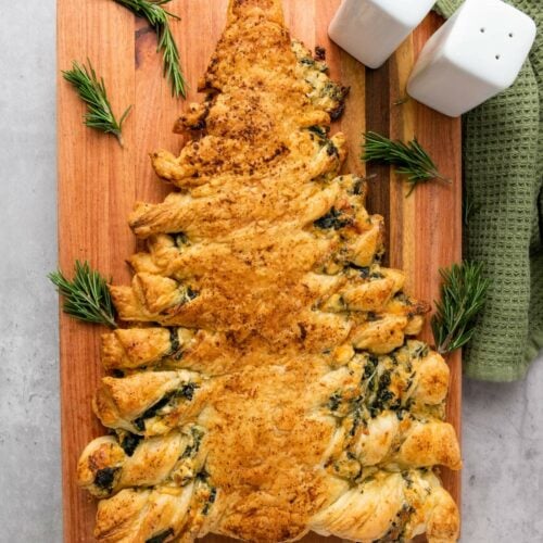 A puff pastry shaped like a Christmas tree, filled with spinach and cheese, sits on a wooden board, garnished with rosemary sprigs.