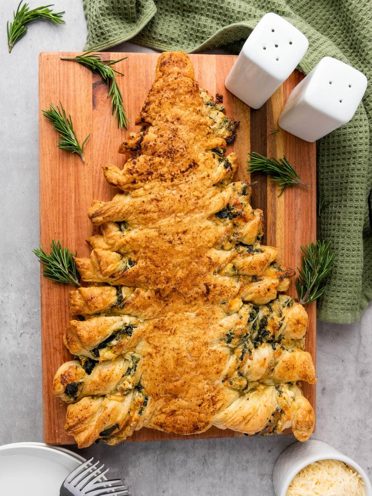 A puff pastry shaped like a Christmas tree, filled with spinach and cheese, sits on a wooden board, garnished with rosemary sprigs.