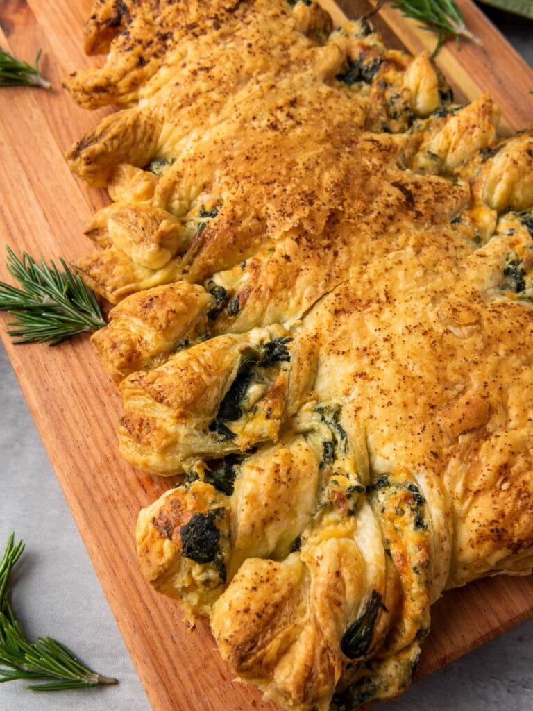 A golden-brown puff pastry filled with spinach and cheese sits on a wooden board, garnished with sprigs of fresh rosemary.