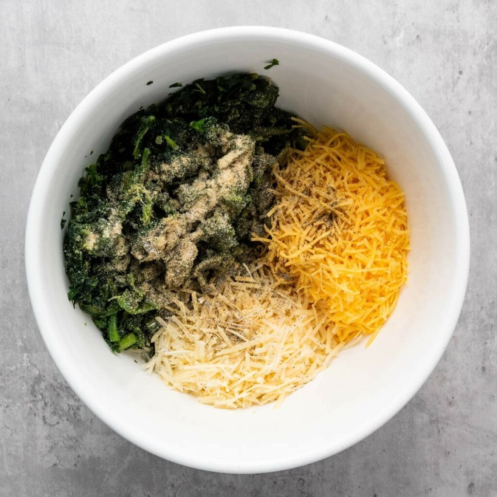 White bowl containing chopped spinach with seasonings, shredded yellow cheese, and shredded parmesan cheese on a gray surface.