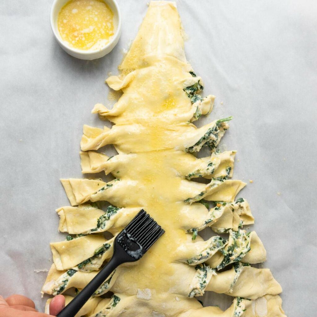 Unbaked puff pastry shaped like a Christmas tree, filled with spinach mixture, is being brushed with butter using a black pastry brush.