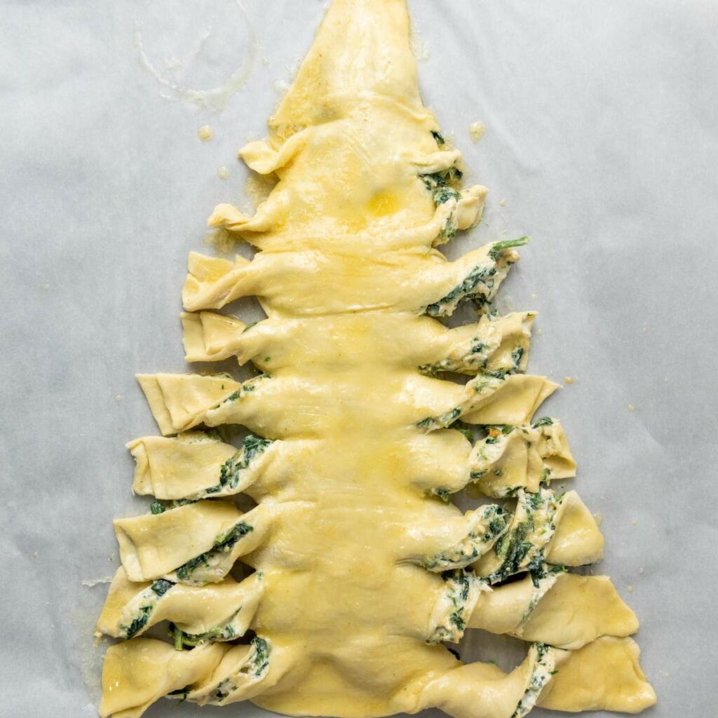 Unbaked puff pastry shaped like a Christmas tree, filled with a spinach mixture and twisted branches, on parchment paper.