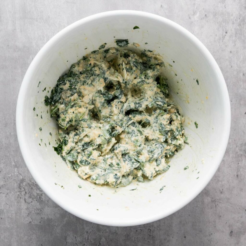 A white bowl containing a mixture of chopped spinach, cheese, and other ingredients on a light gray surface.