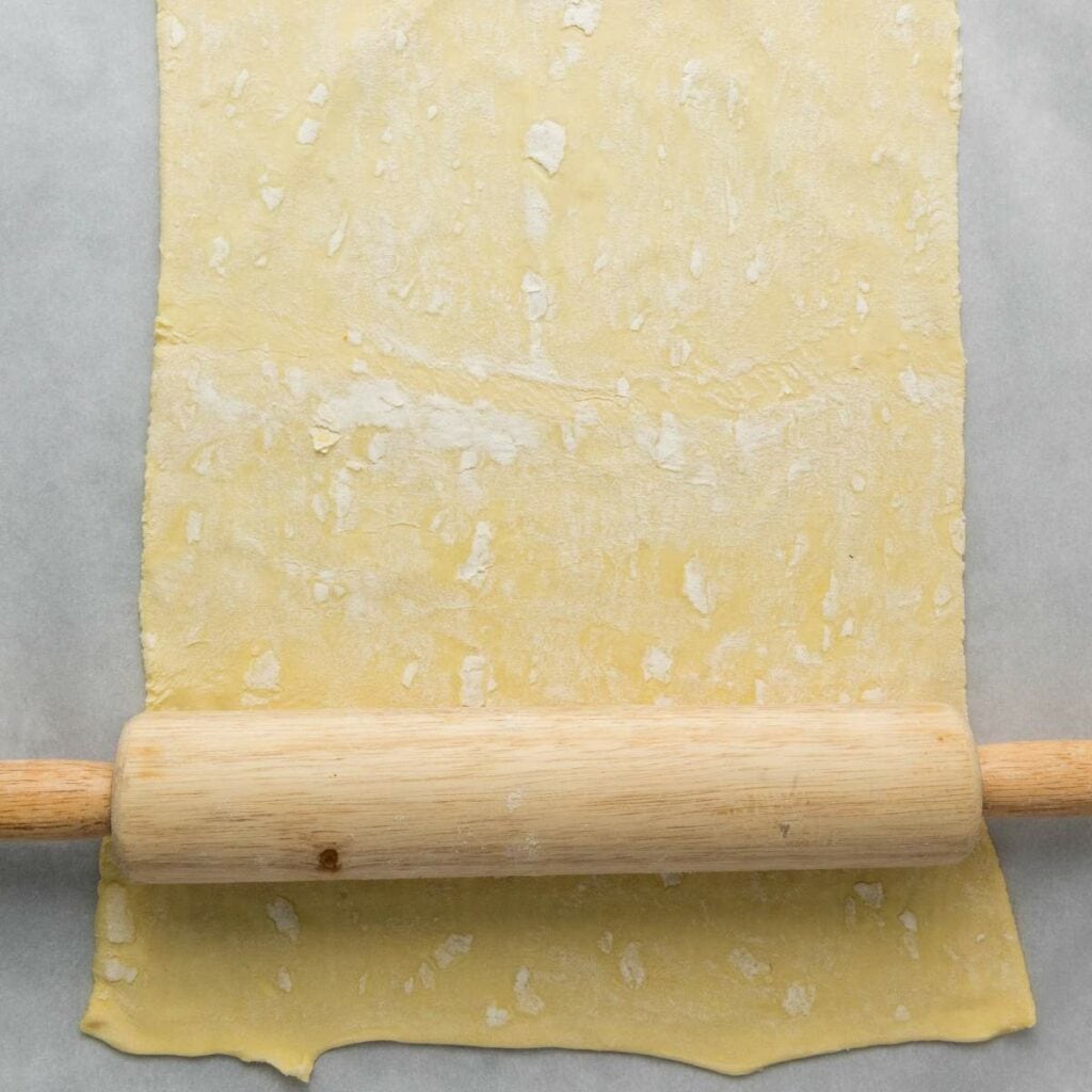 A wooden rolling pin rolling out a sheet of dough on parchment paper.
