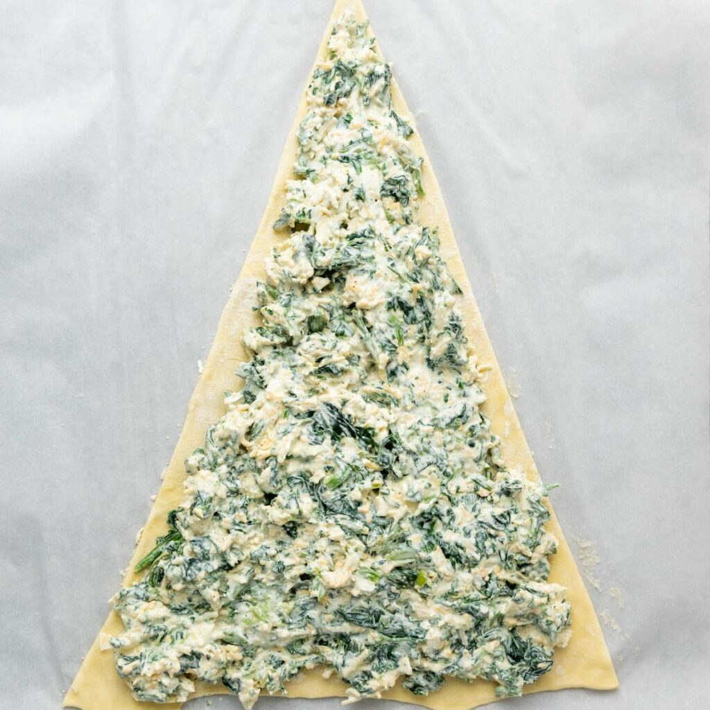 A large triangle of dough topped with a spinach and cheese mixture, arranged on a sheet of parchment paper.