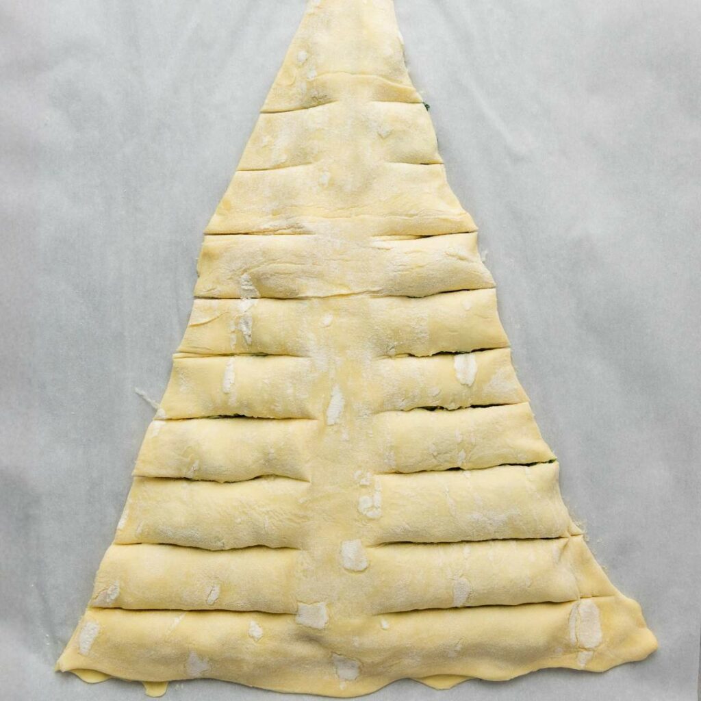 Triangle-shaped sheet of dough with horizontal slits cut on both sides, placed on parchment paper.