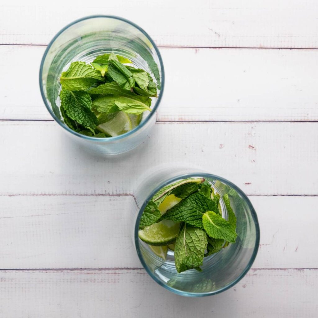 Two clear glasses containing fresh mint leaves and lime wedges on a white wooden surface, viewed from above.