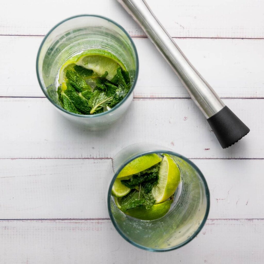 Two glasses with lime wedges and mint leaves on a white wooden surface, next to a metal muddler.