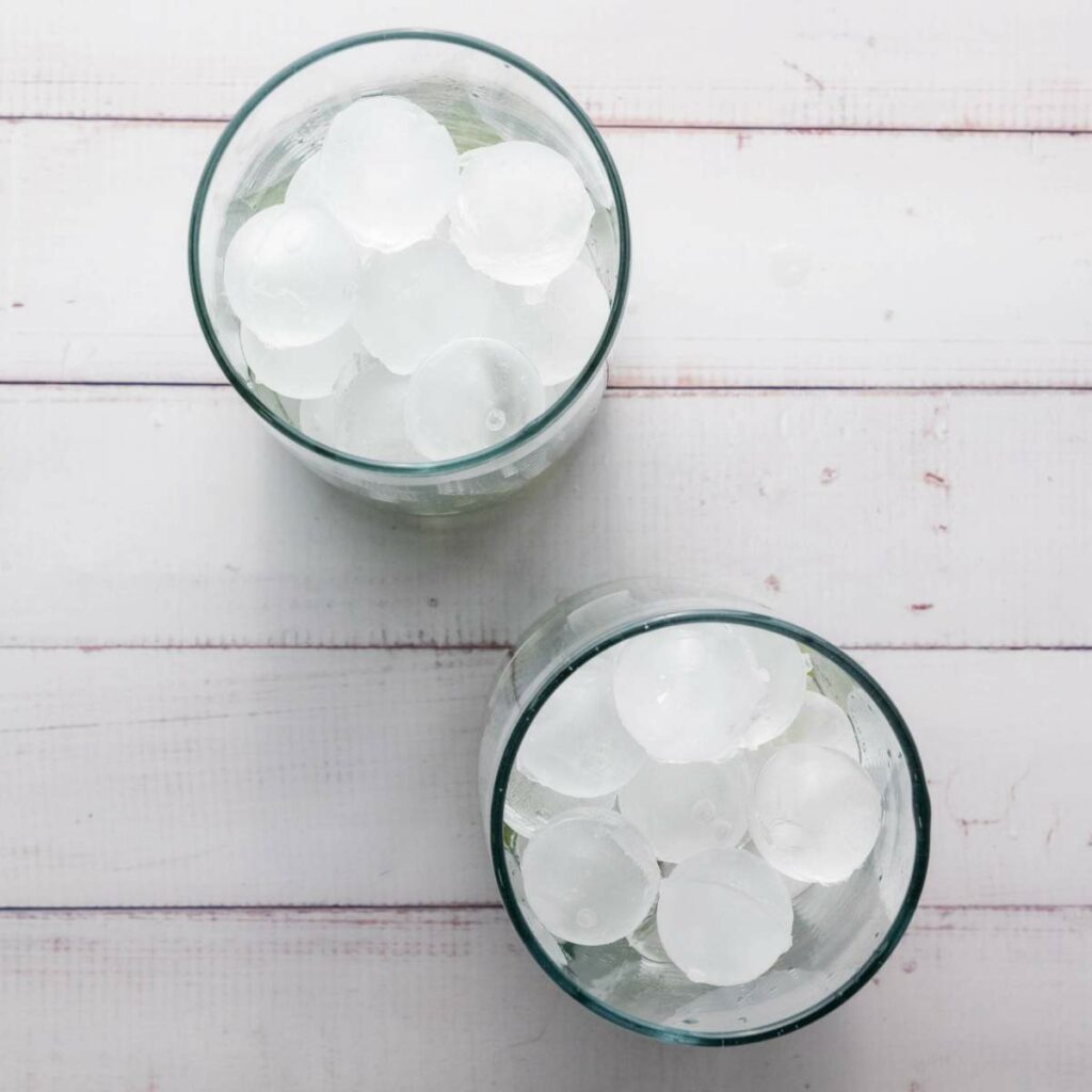 Two glasses filled with round ice cubes on a white wooden surface, viewed from above.