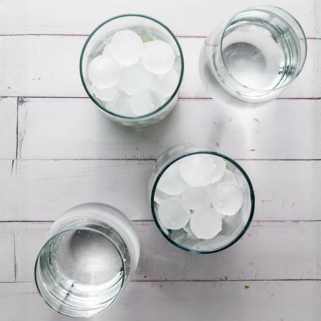 Four clear glasses of water, two with ice cubes and two without, are arranged on a white wooden surface viewed from above.