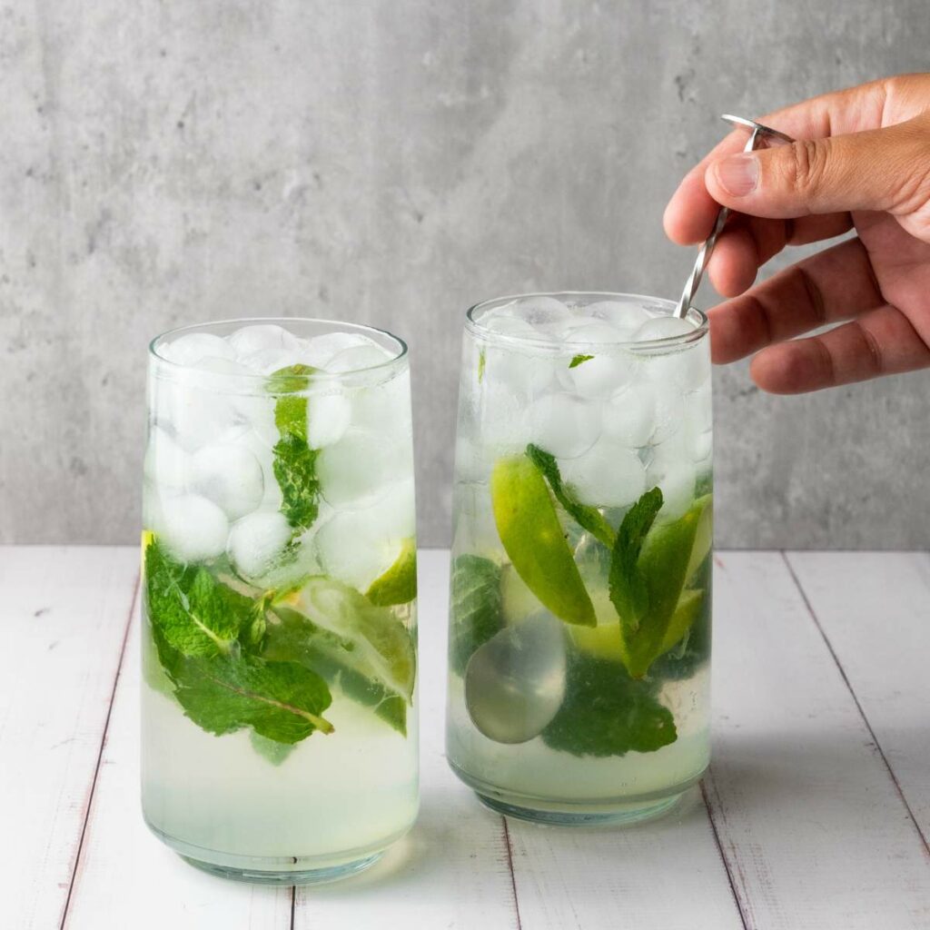 Two glasses of iced mojito with lime wedges and mint leaves; a hand is stirring one of the drinks with a metal spoon.
