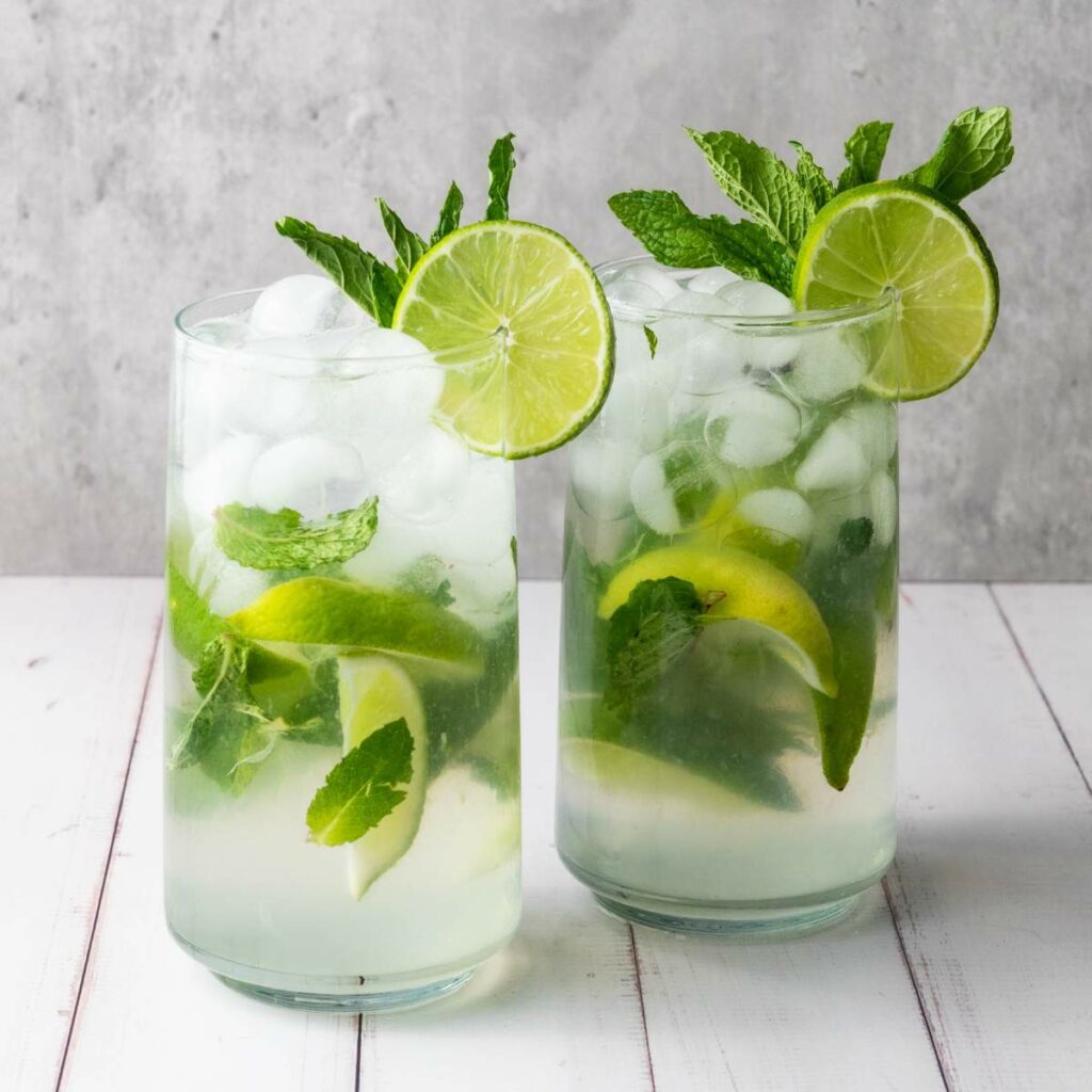 Two glasses of mojito with ice, lime slices, and fresh mint on a white surface against a light gray background.