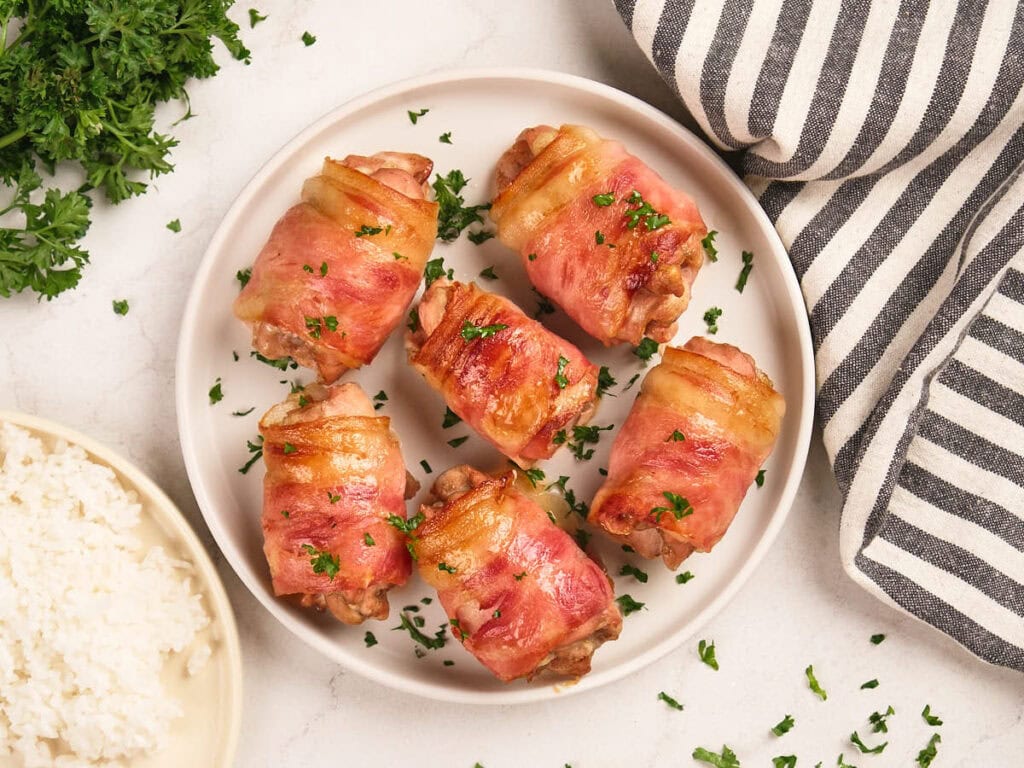A plate with six bacon-wrapped chicken pieces garnished with chopped parsley, next to a bowl of white rice and a striped cloth napkin.