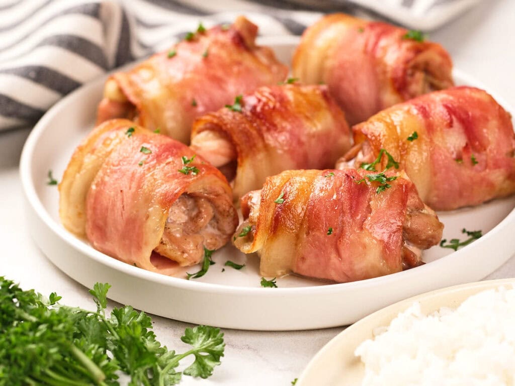 A plate of bacon-wrapped chicken pieces garnished with chopped parsley, with a side of rice and fresh parsley visible nearby.