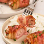 A fork holds a piece of bacon-wrapped chicken above a plate with more pieces of bacon-wrapped chicken and a serving of white rice, garnished with chopped parsley.