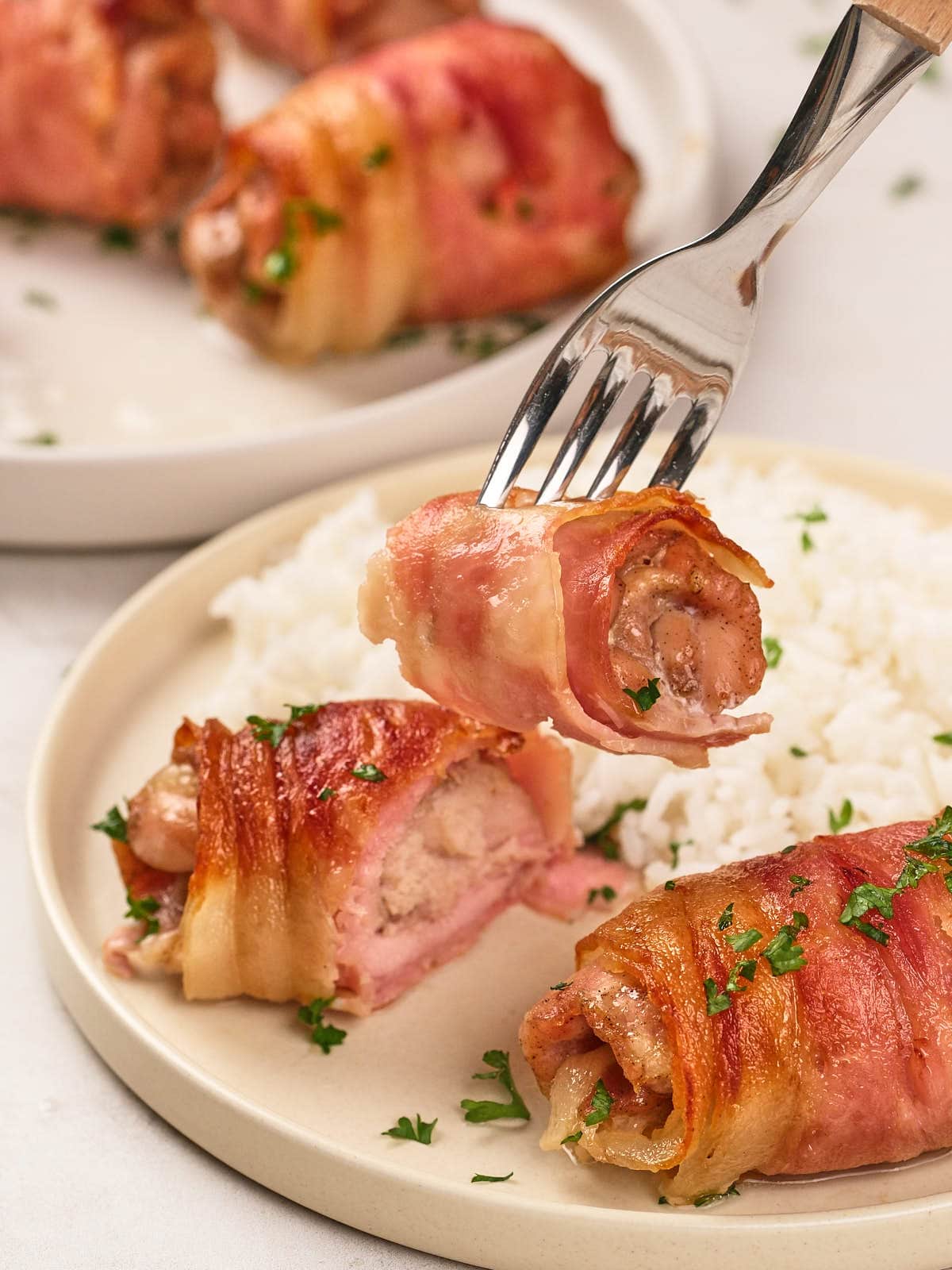 A fork holds a piece of bacon-wrapped chicken above a plate with more pieces of bacon-wrapped chicken and a serving of white rice, garnished with chopped parsley.
