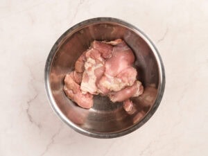 Raw pieces of chicken in a metal mixing bowl on a light-colored marble surface.