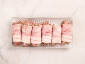 Six pieces of raw bacon-wrapped meat arranged in a row on a rectangular white plate, set on a light marble surface.
