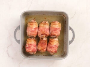 Six bacon-wrapped chicken thighs arranged in a square metal baking dish on a light marble surface.