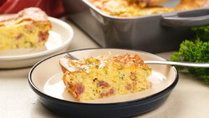 A slice of baked omelet with visible pieces of ham and herbs on a plate, with the remaining casserole in a baking dish in the background.