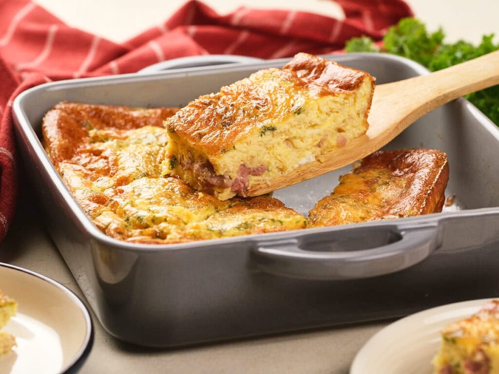 A baked egg casserole with visible pieces of ham and herbs is being served from a square pan with a wooden spatula.