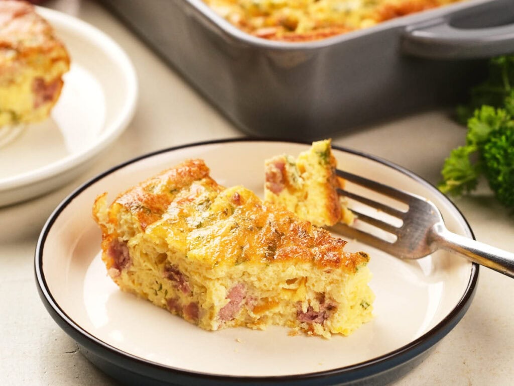A slice of baked omelet with ham and cheese on a plate, with a fork and a casserole dish in the background.