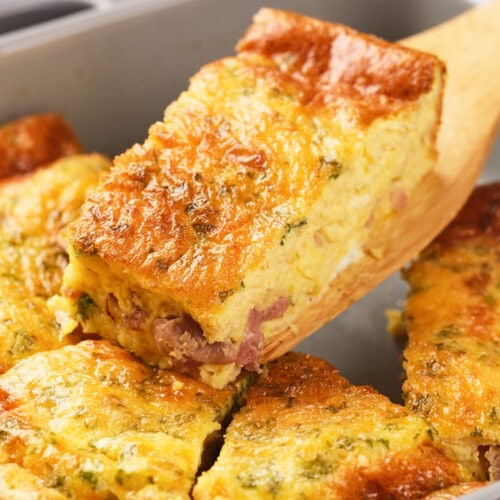 A wooden spatula lifts a slice of baked omelet with visible ham and herbs from a square baking dish.
