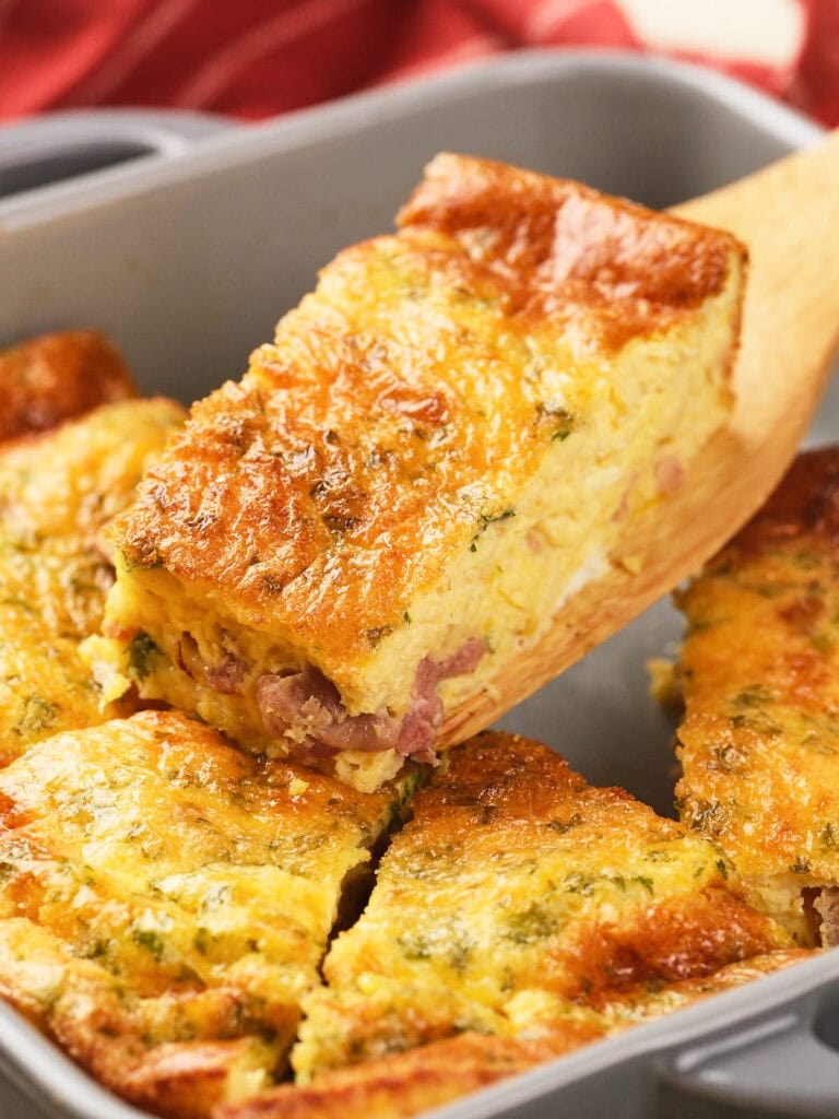 A wooden spatula lifts a slice of baked omelet with visible ham and herbs from a square baking dish.