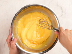 A person is whisking a thick yellow mixture in a stainless steel bowl on a light-colored countertop.