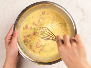 A person whisking a mixture of eggs, chopped vegetables, and meat in a stainless steel bowl on a light countertop.