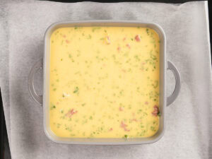A square baking dish filled with an uncooked egg mixture containing herbs and pieces of meat, placed on parchment paper.