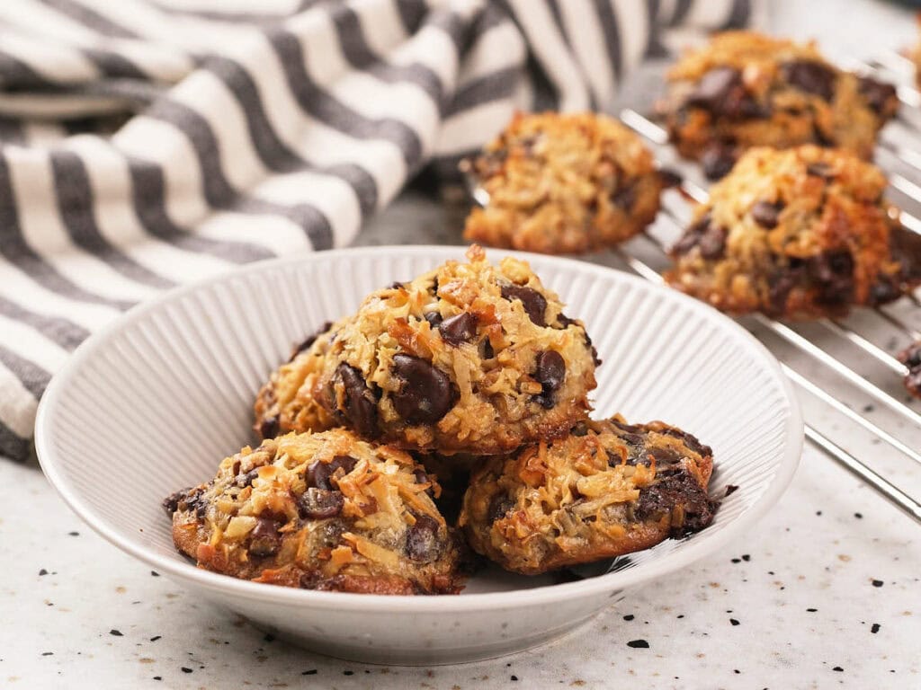 A white bowl holds several magic cookies, with more cookies cooling on a wire rack in the background and a striped cloth nearby.