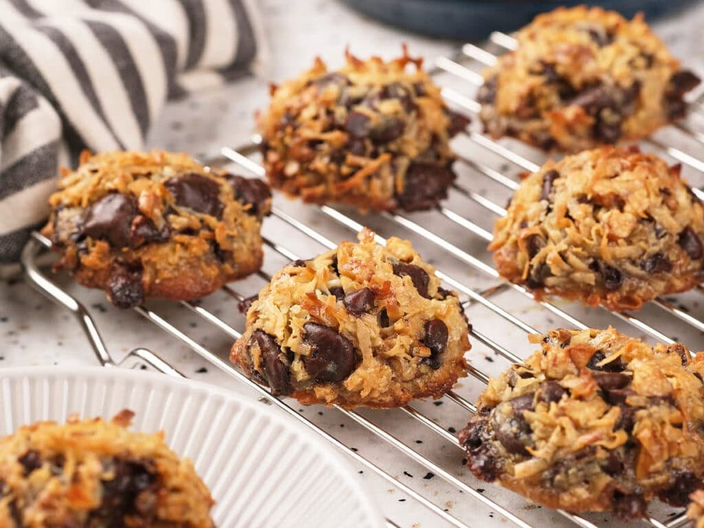 A cooling rack with several chocolate chip coconut cookies, with a striped cloth and a white plate in the background.