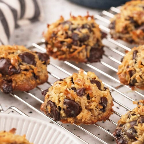Several coconut and chocolate chip cookies cooling on a wire rack, with a striped cloth and more cookies visible in the background.