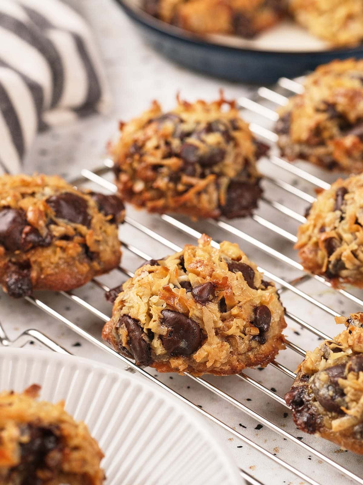 Several coconut and chocolate chip cookies cooling on a wire rack, with a striped cloth and more cookies visible in the background.