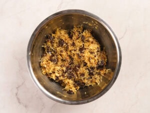 A stainless steel bowl containing a mixture of shredded coconut, raisins, and other baking ingredients on a light-colored countertop.