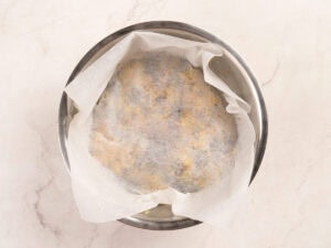 A metal bowl lined with parchment paper contains dough, partially covered by the paper, on a light-colored countertop.