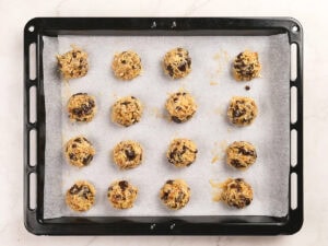 A baking tray lined with parchment paper holds sixteen unbaked cookie dough balls with chocolate chips, spaced evenly in rows.