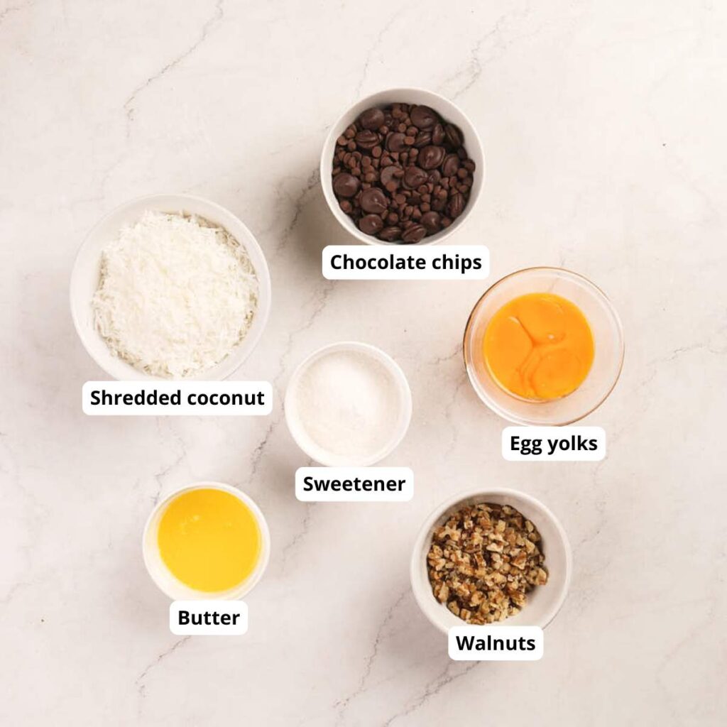 Six bowls on a marble surface contain chocolate chips, egg yolks, shredded coconut, sweetener, melted butter, and chopped walnuts, each labeled with text.