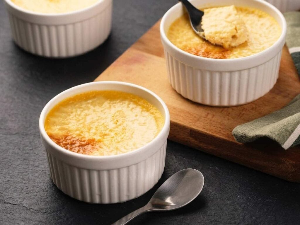Three ramekins of baked custard are arranged on a dark surface, with one partially eaten and a spoon resting nearby.