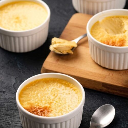 Four white ramekins filled with baked custard are arranged on a dark surface, with a spoonful of custard on a wooden board and an empty spoon beside one ramekin.