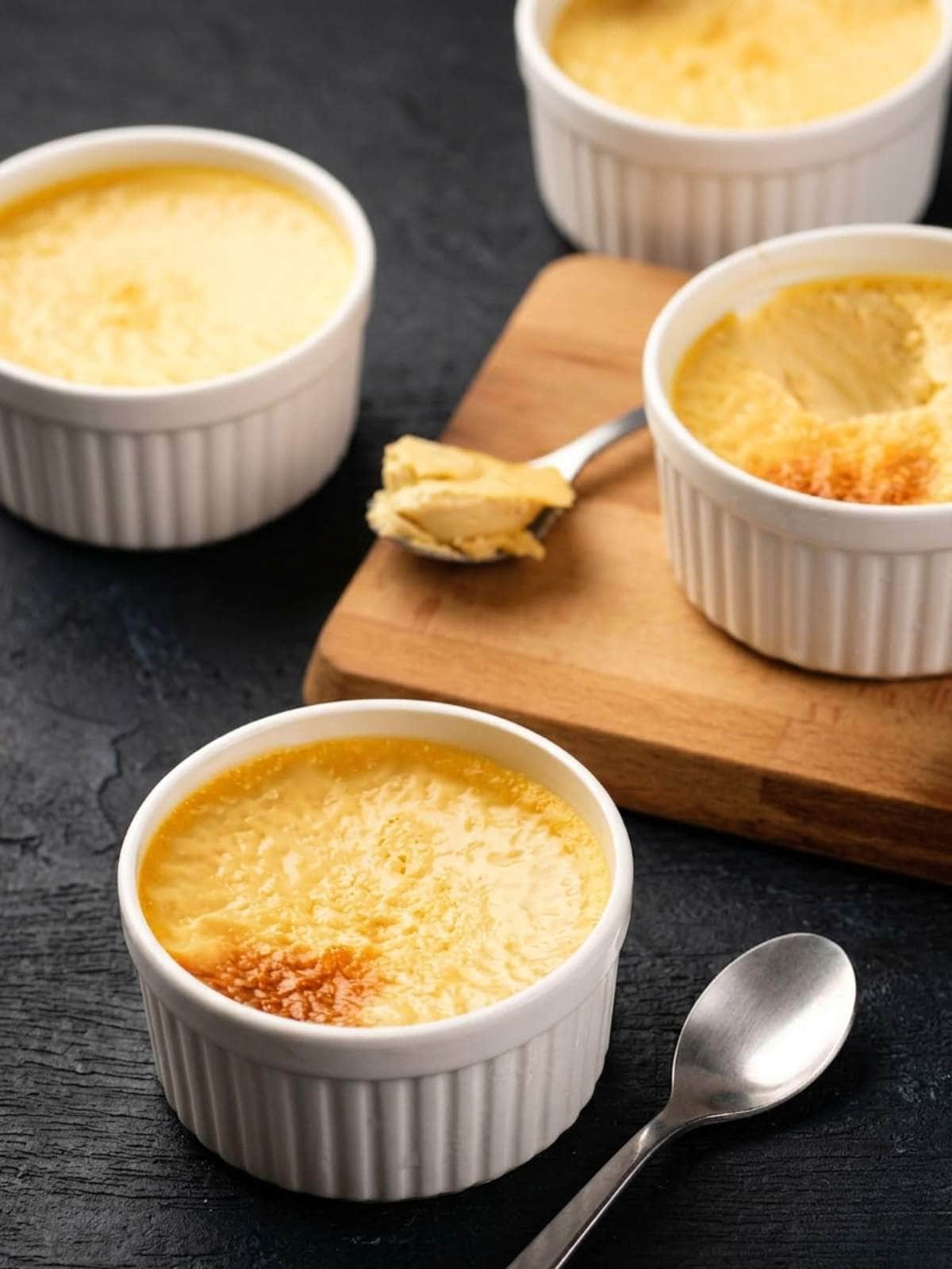 Four white ramekins filled with baked custard are arranged on a dark surface, with a spoonful of custard on a wooden board and an empty spoon beside one ramekin.