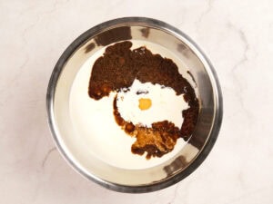 A metal mixing bowl containing cream, brown sugar, vanilla extract, and an egg on a light countertop.