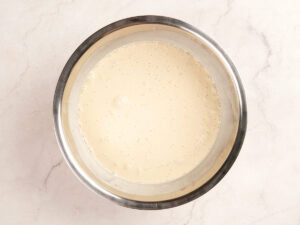 A stainless steel bowl filled with a smooth, pale yellow batter sits on a light-colored marble surface.