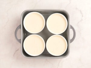 Four white ramekins filled with a light cream mixture are arranged inside a gray baking dish on a light marble surface.